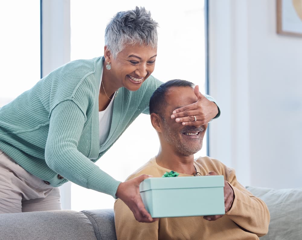 An older woman surprises a man with a gift box, playfully covering his eyes with her hand in a cozy living room setting, capturing a moment of joy and surprise between them.