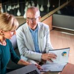 A senior woman and man are reviewing financial documents and a colorful pie chart on a laptop screen, likely discussing investment strategies in a modern office setting.