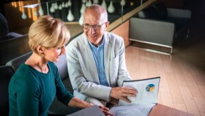 A senior woman and man are reviewing financial documents and a colorful pie chart on a laptop screen, likely discussing investment strategies in a modern office setting.