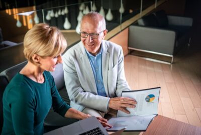 A senior woman and man are reviewing financial documents and a colorful pie chart on a laptop screen, likely discussing investment strategies in a modern office setting.