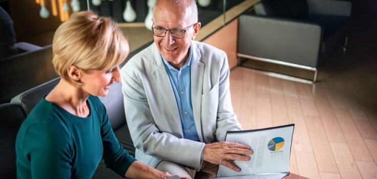 A senior woman and man are reviewing financial documents and a colorful pie chart on a laptop screen, likely discussing investment strategies in a modern office setting.