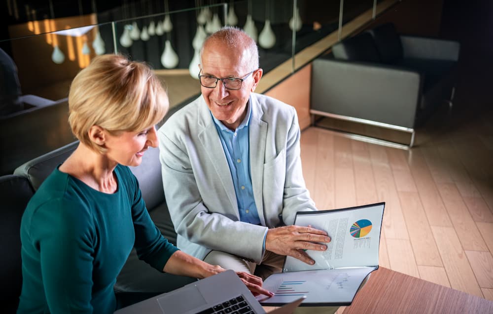 A senior woman and man are reviewing financial documents and a colorful pie chart on a laptop screen, likely discussing investment strategies in a modern office setting.